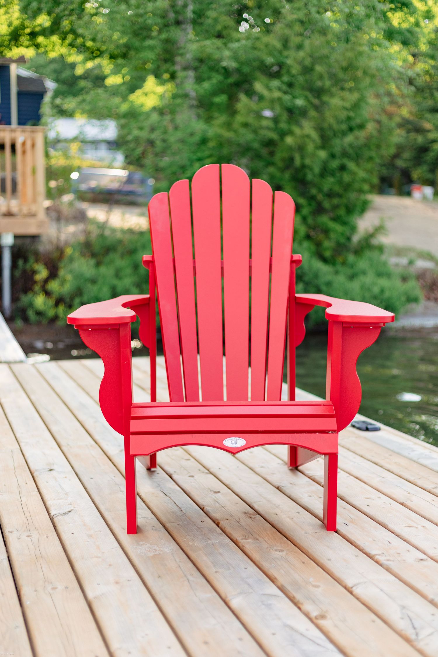 A bright red Adirondack chair sits on a wooden deck overlooking a lake with green foliage in the background.
