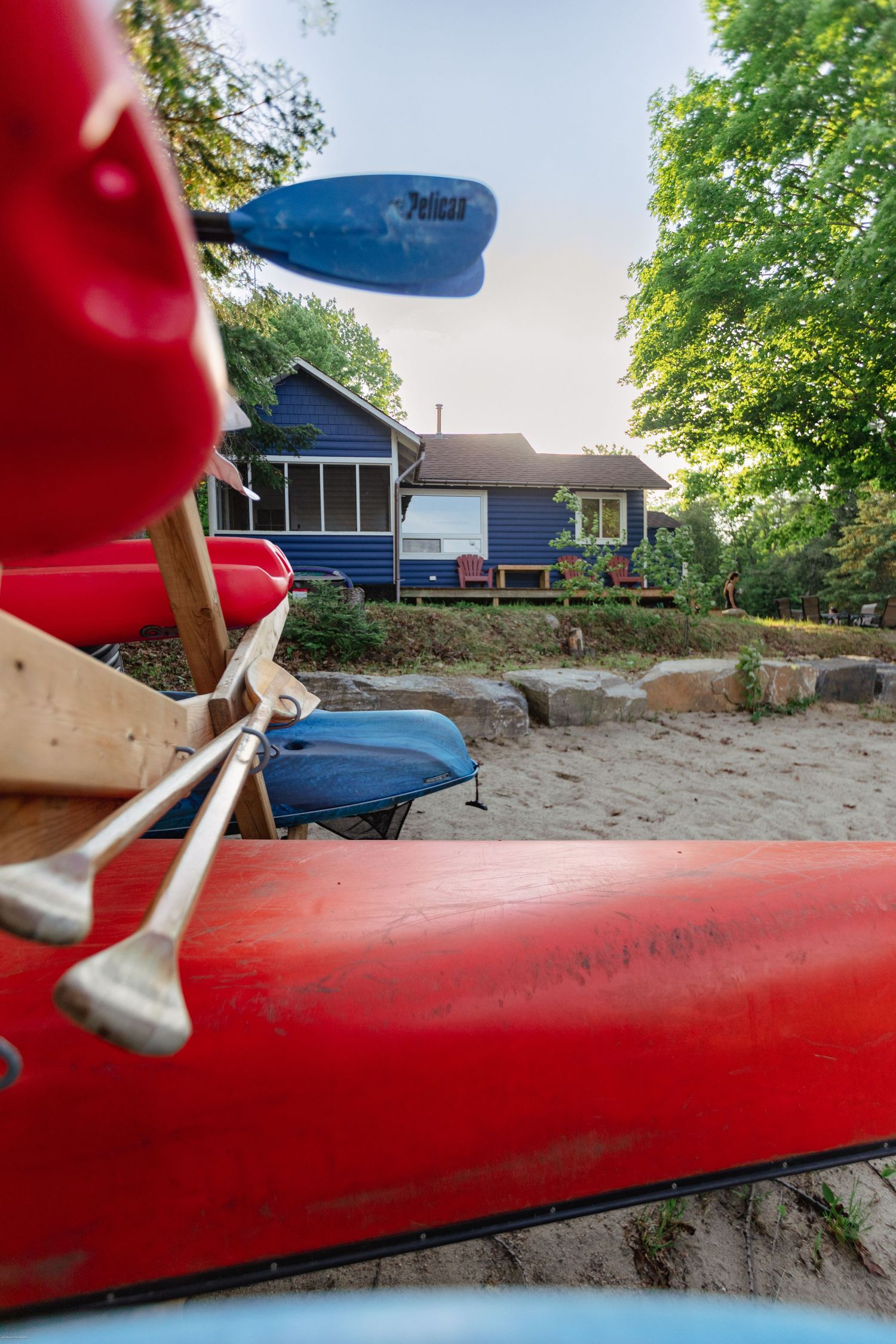A red kayak with a blue paddle rests on a beach with a lake house visible in the distance.