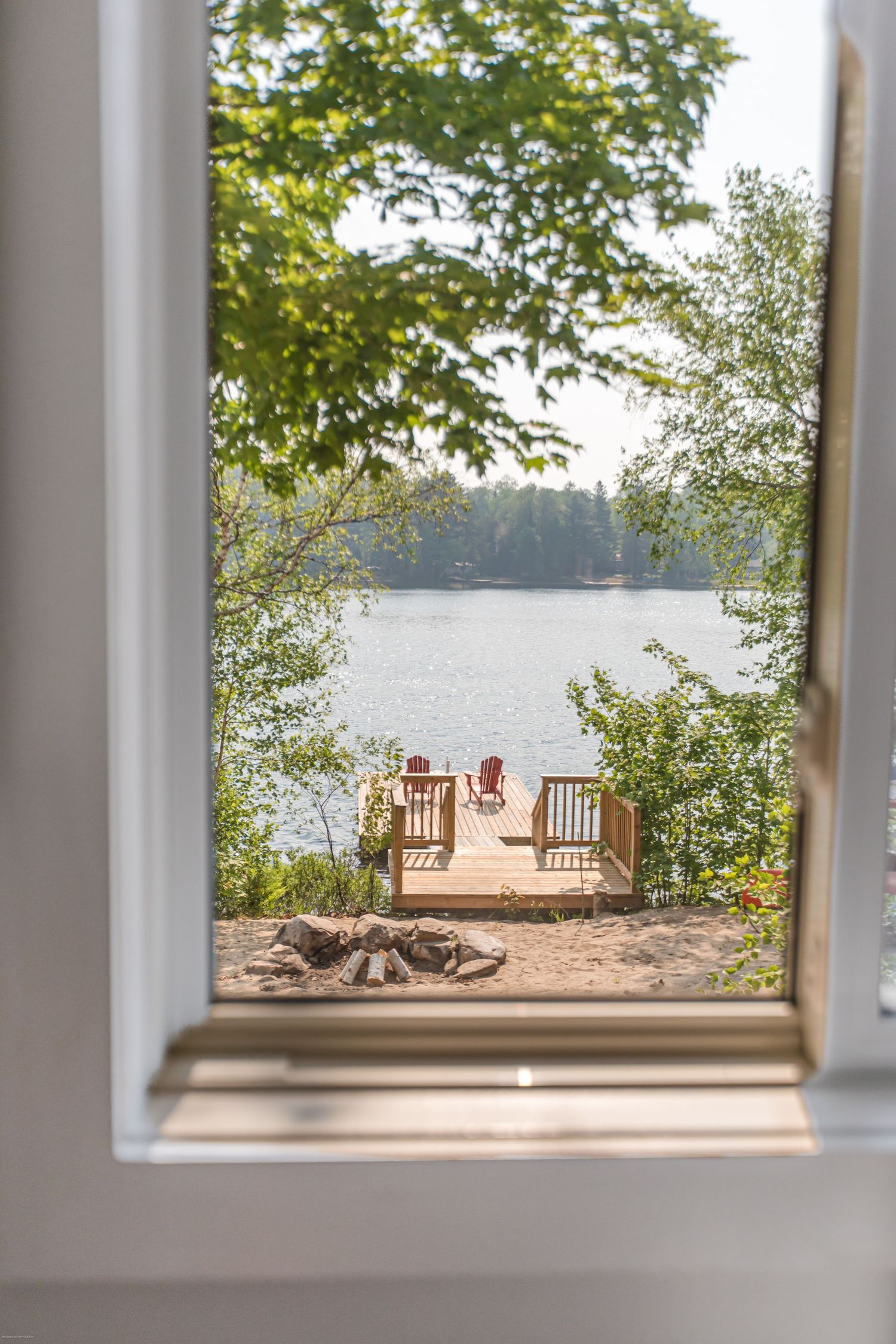 Window view of a lakeside dock with Adirondack chairs framed by trees and natural scenery.