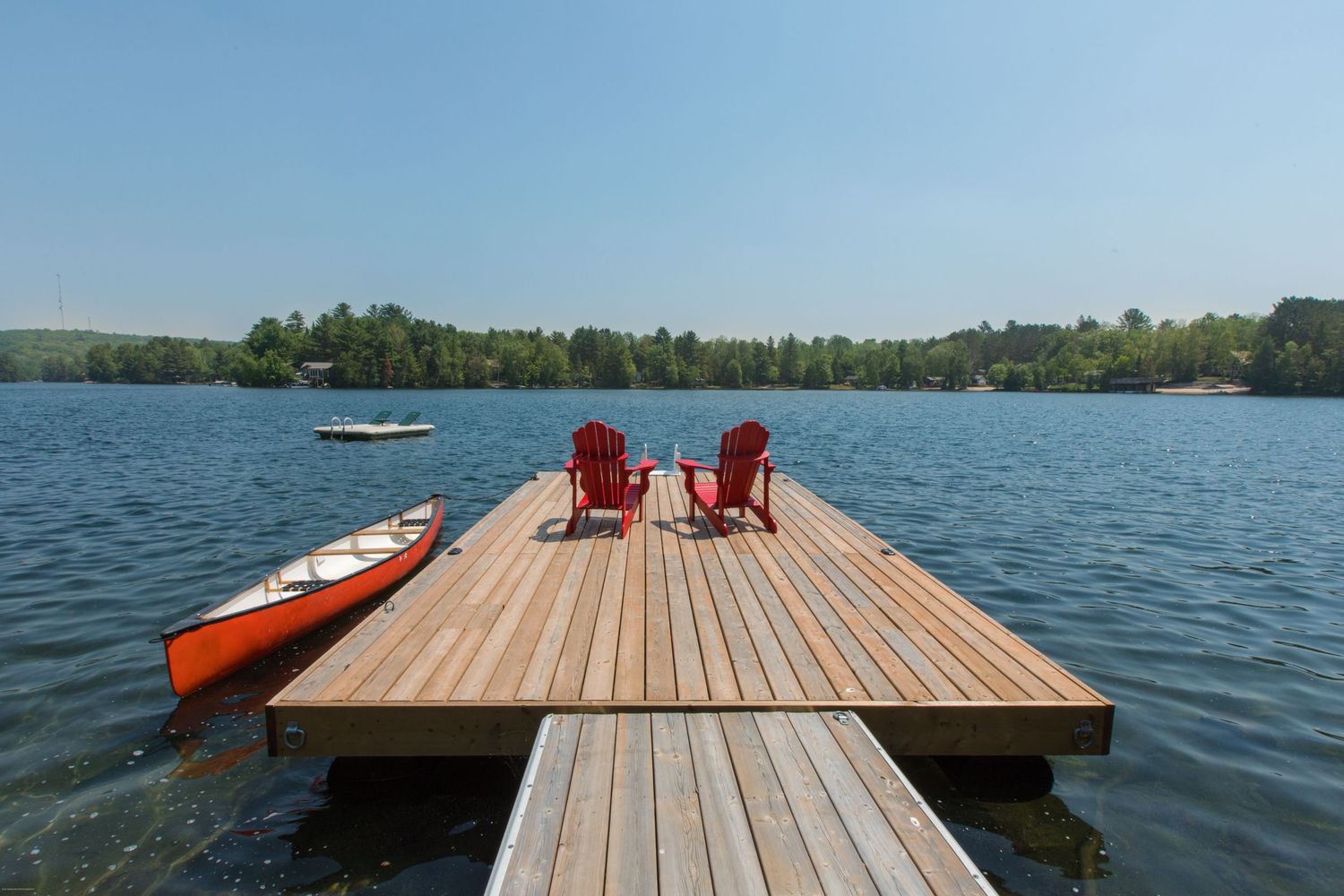 Two red Adirondack chairs sit on a wooden dock with orange kayaks beside them overlooking a serene lake.