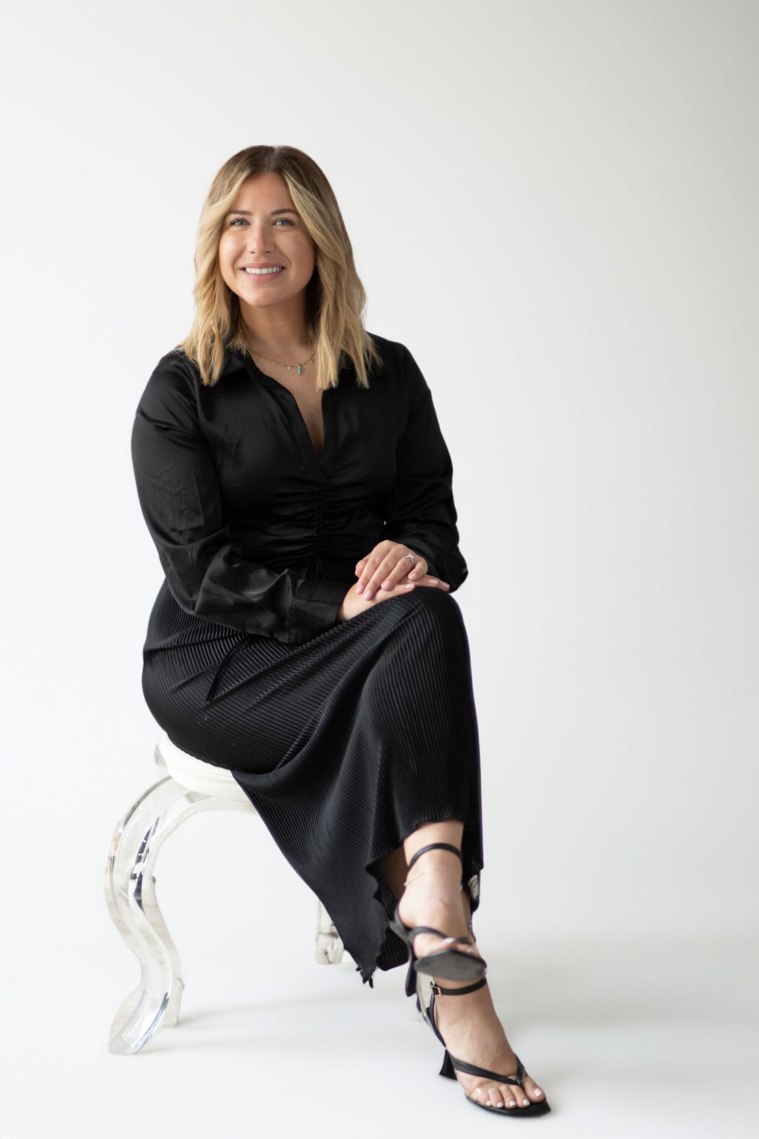 Woman in black dress sitting on clear acrylic chair against white background.