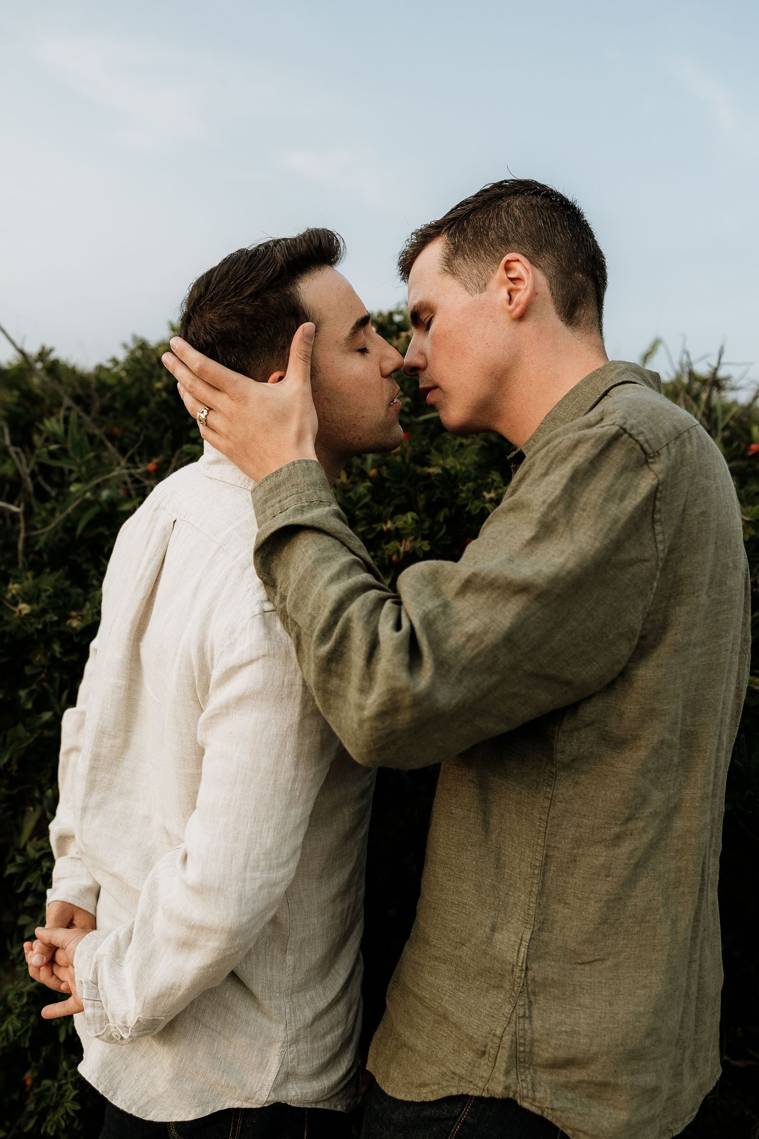 Two men sharing an intimate moment, one gently cupping the other's face as they lean in for a kiss outdoors. Westport Ma. Miranda Photo & Co