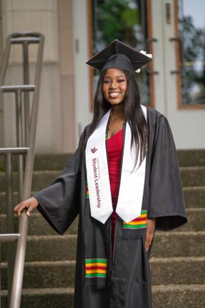 Graduate in red dress and black gown stands proudly on campus steps.