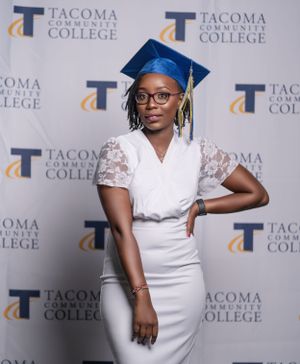 Graduate poses in white dress and blue cap against Tacoma Community College backdrop.
