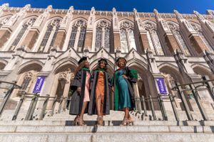 Three graduates in colorful dresses pose on stone steps of Gothic university building.