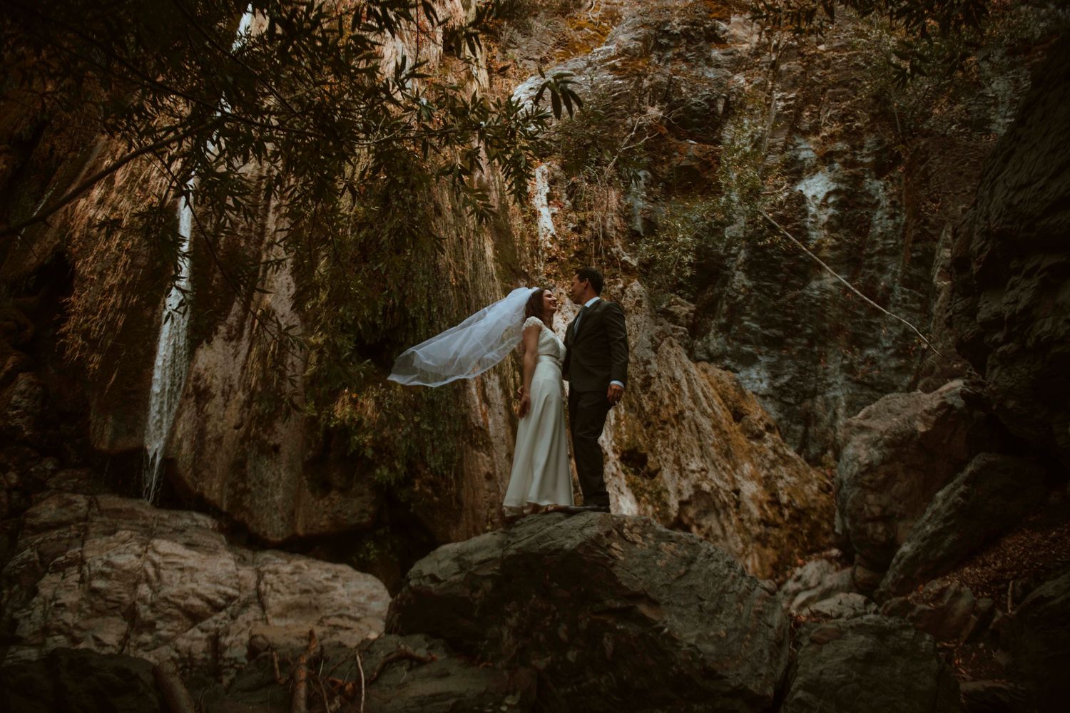 A couple shares a romantic moment under dramatic rock formations in a moody forest setting at dusk.