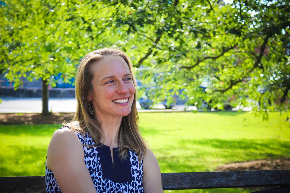 A person smiles joyfully while sitting outdoors against a backdrop of green trees and blue sky.