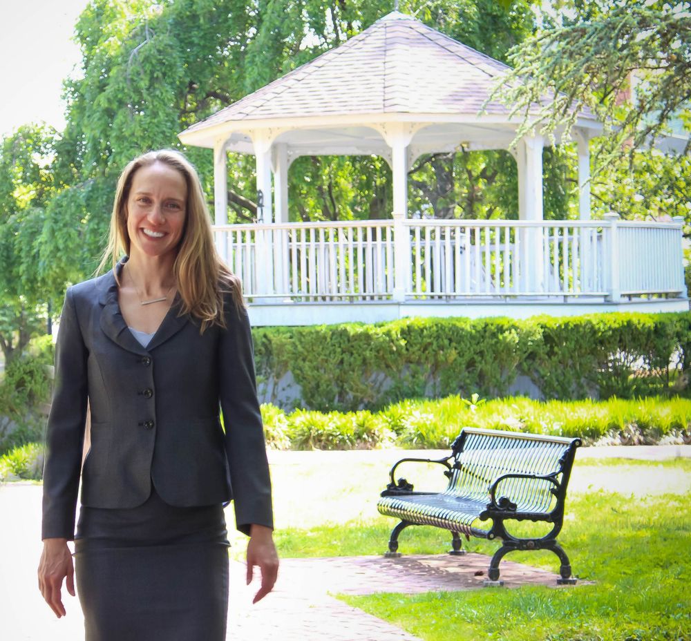 A person in business attire stands near a white gazebo and park bench in a garden setting.