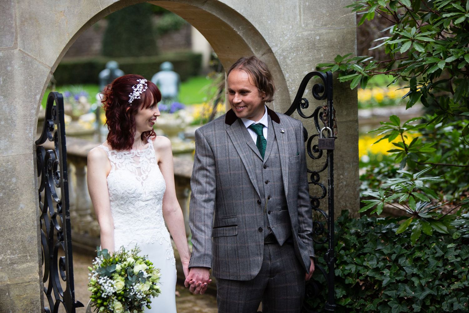 Wedding guests gather in a garden setting with ornate stone architecture and flowering plants.