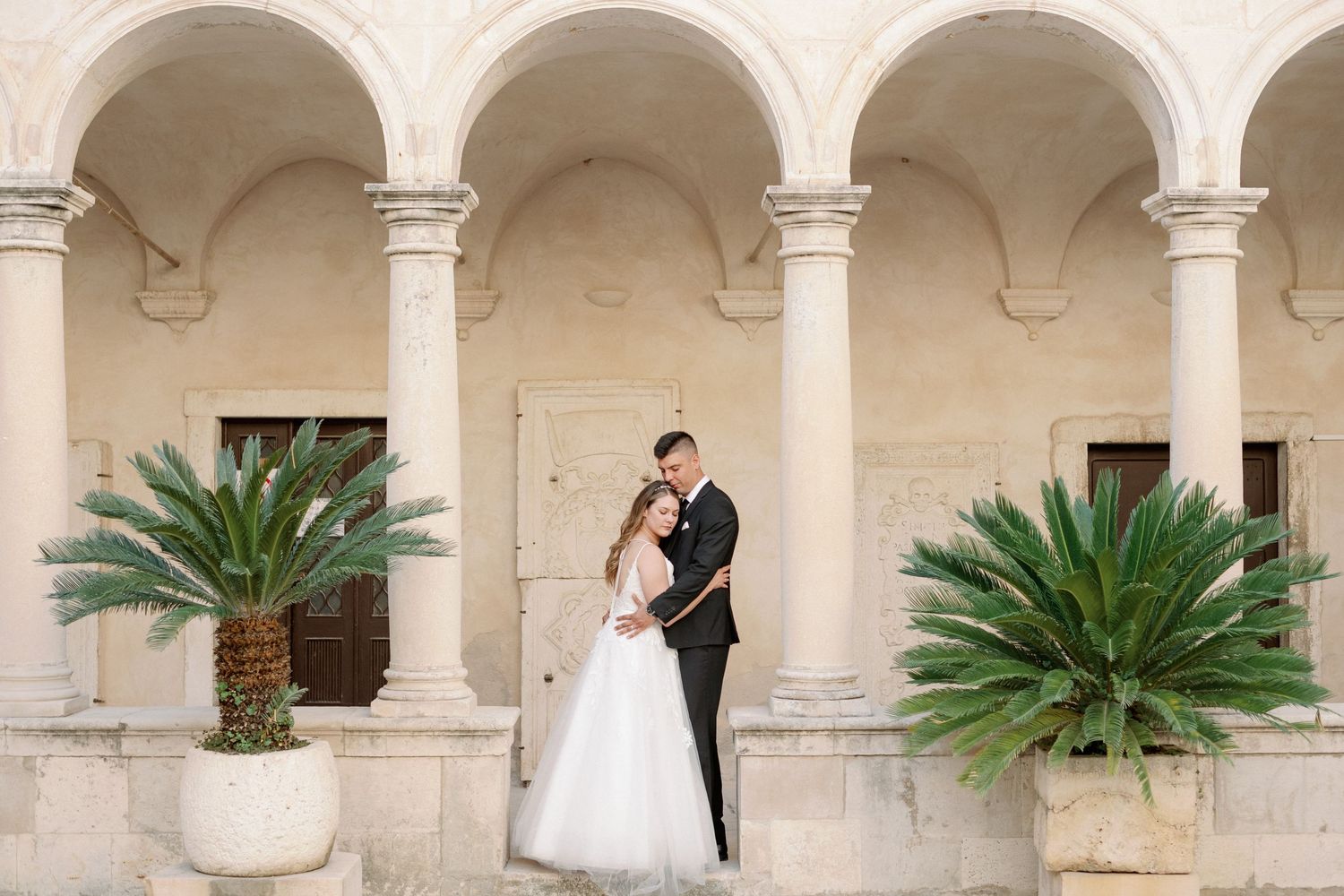 Elegant couple poses beneath white stone archway with decorative palm plants in ornate planters at elegant venue.