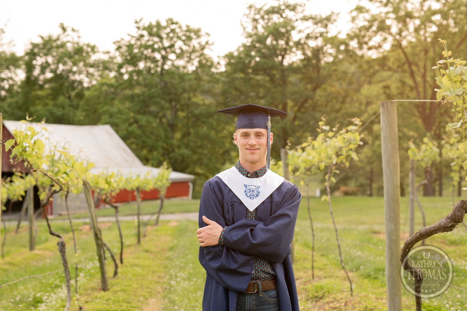 Graduate poses in academic regalia near grapevines with a barn in the background.