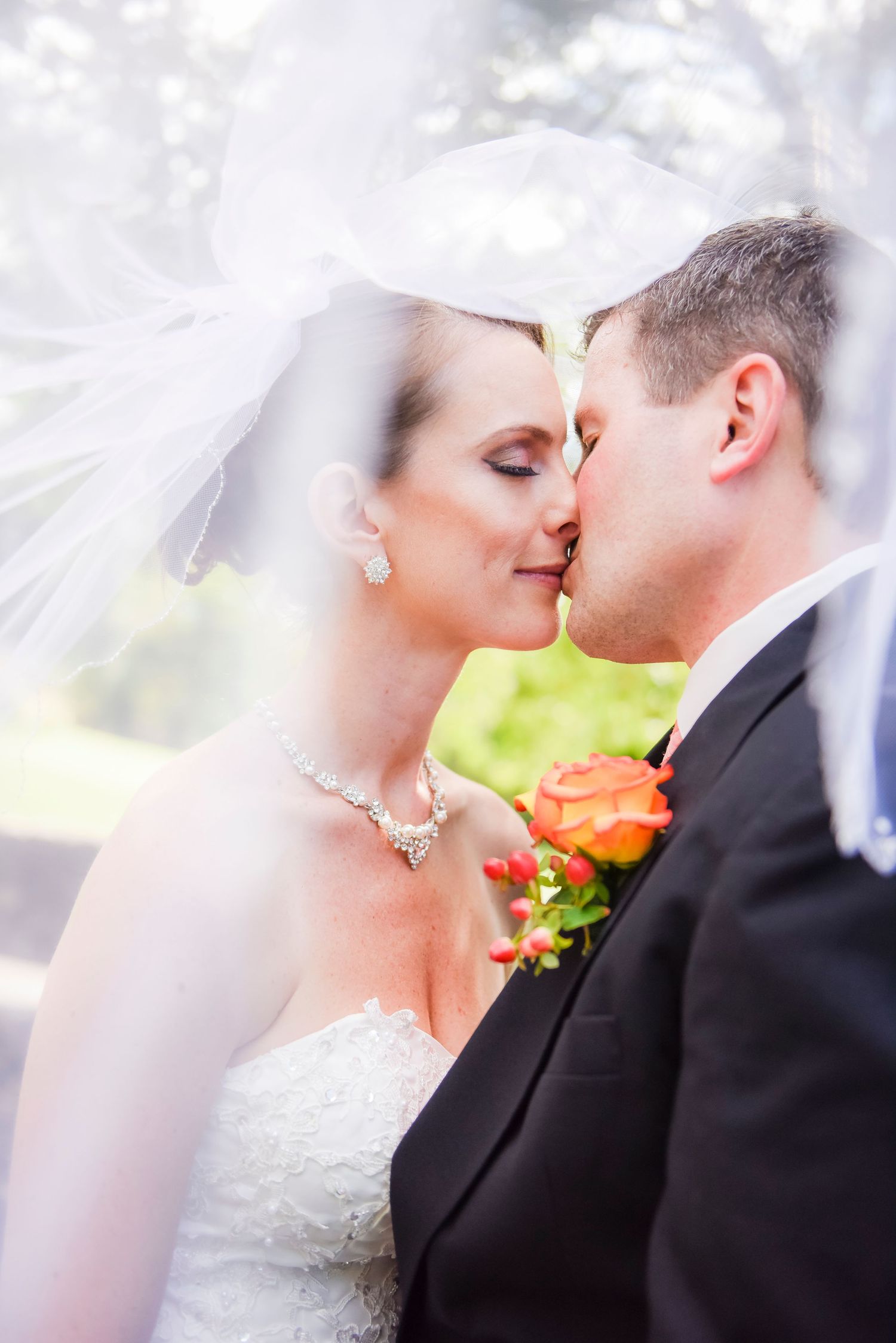 Bride and groom sharing a romantic kiss under flowing white wedding veil outdoors.