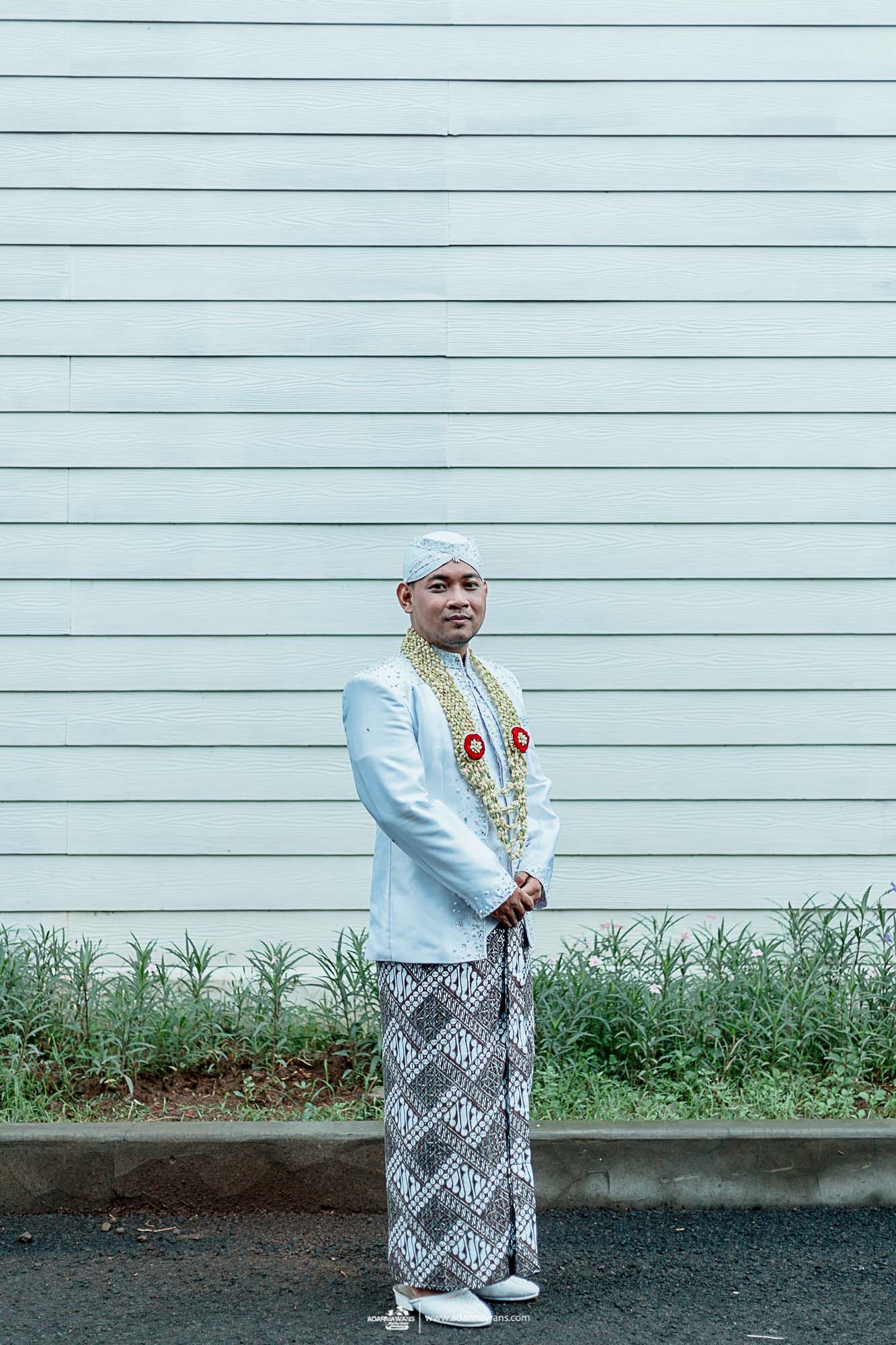 Full length portrait of groom in white ceremonial attire against gray siding.