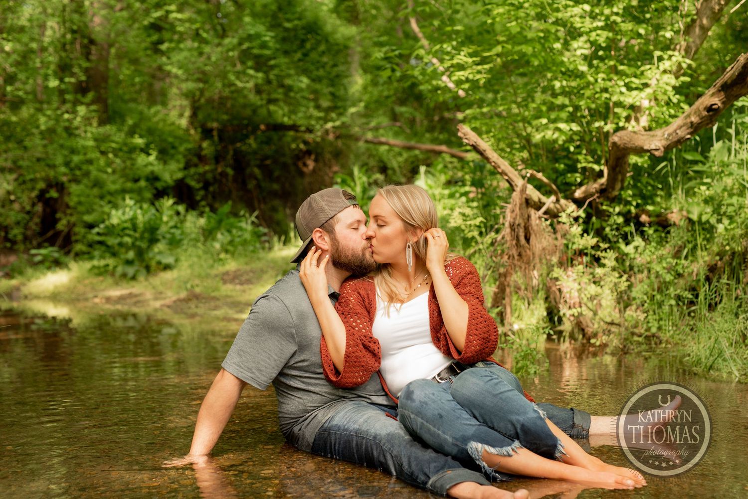 A couple shares a tender moment while sitting in a shallow creek with sunlit forest behind them.