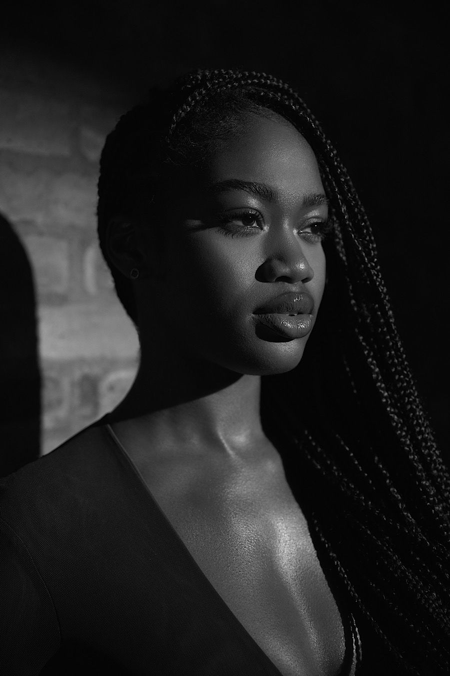 Dramatic black and white portrait showcasing long braided hair against brick wall background.