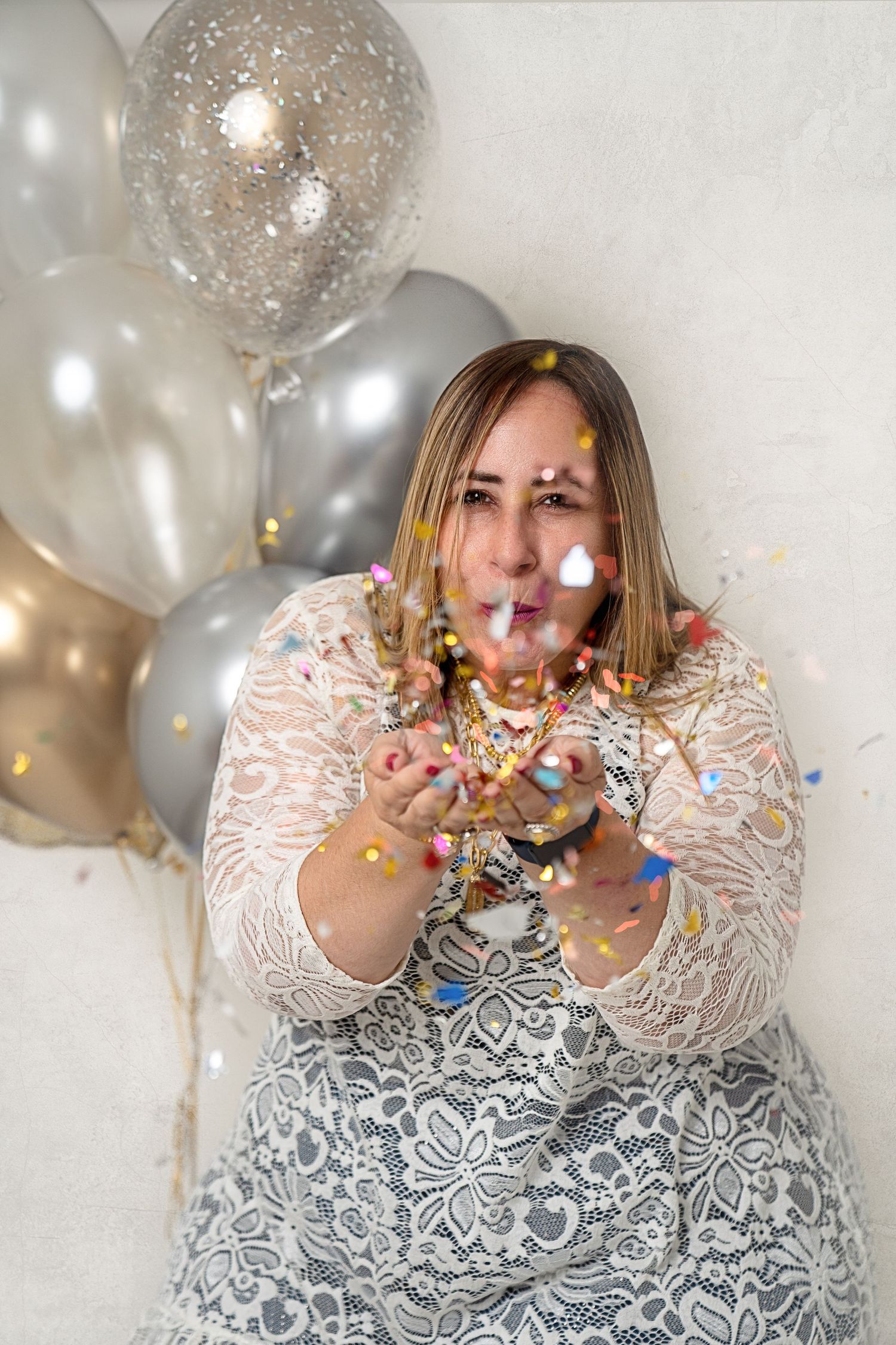 Woman in white lace dress blowing glittery confetti surrounded by silver and gold balloons.