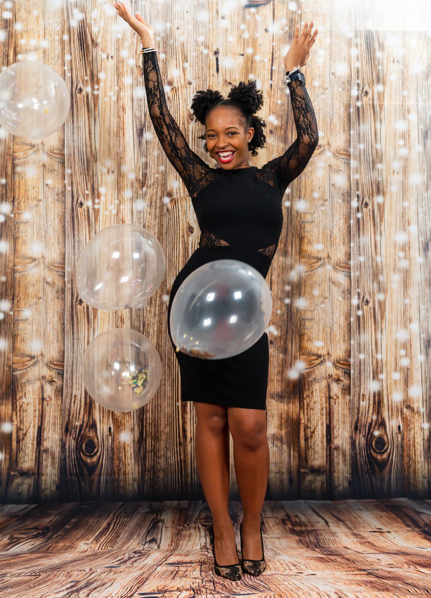 Happy woman in black lace dress celebrating with clear balloons against rustic wood background.