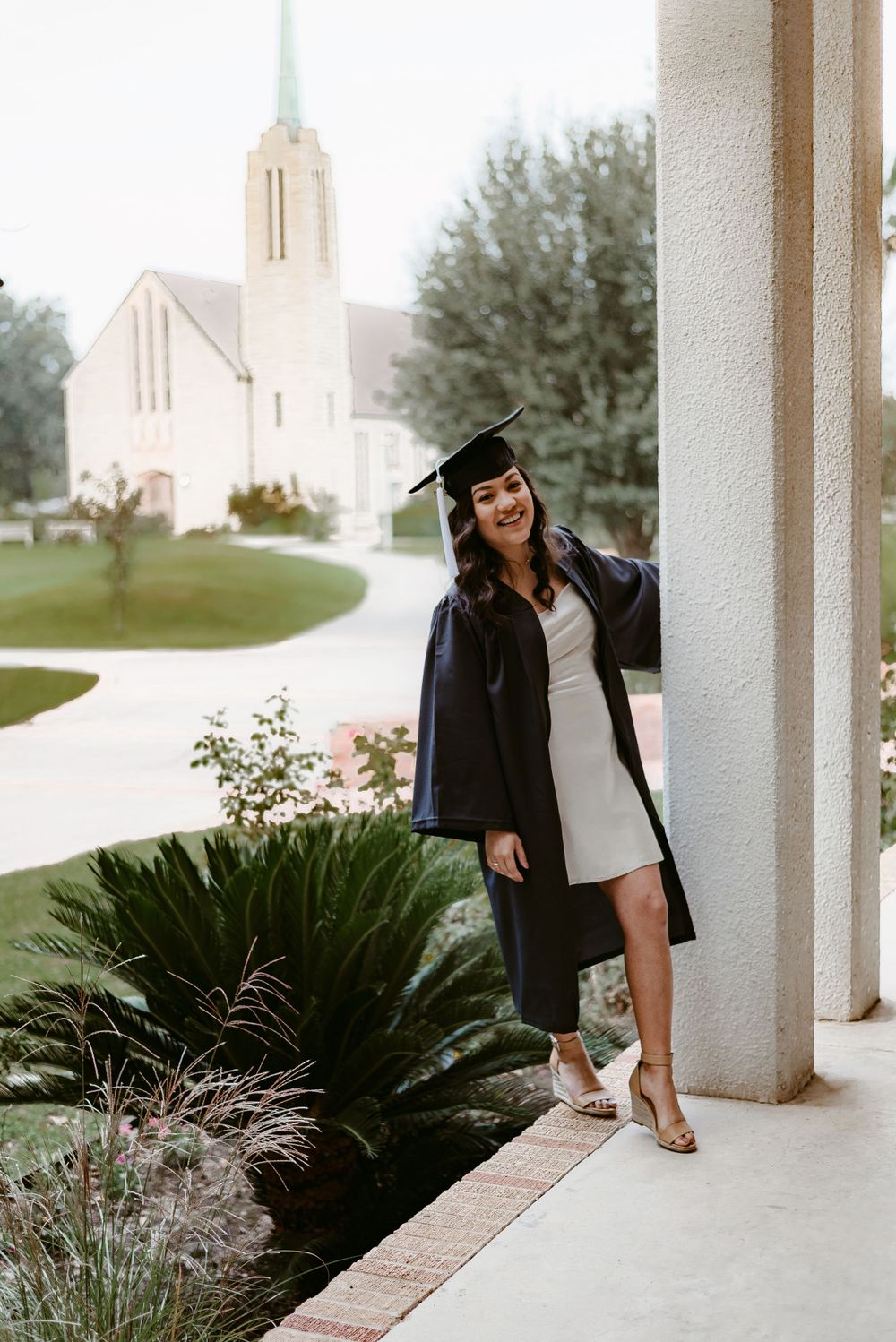Graduate in cap and gown stands on pathway with white church building visible in background.