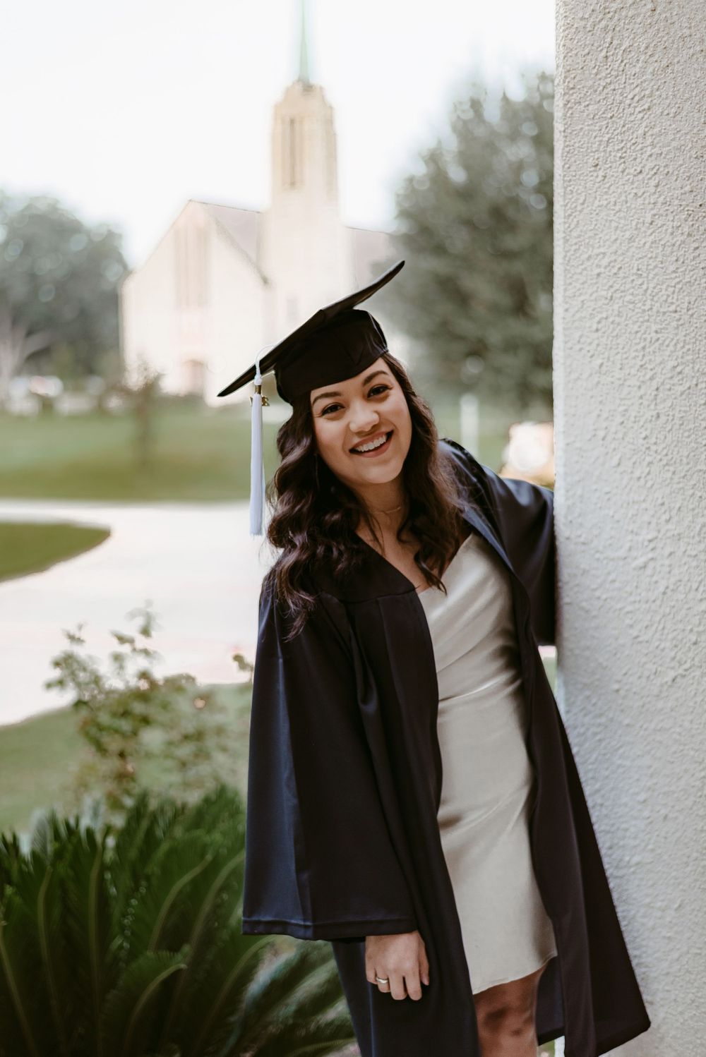 Graduate in academic regalia poses near white church building with steeple.