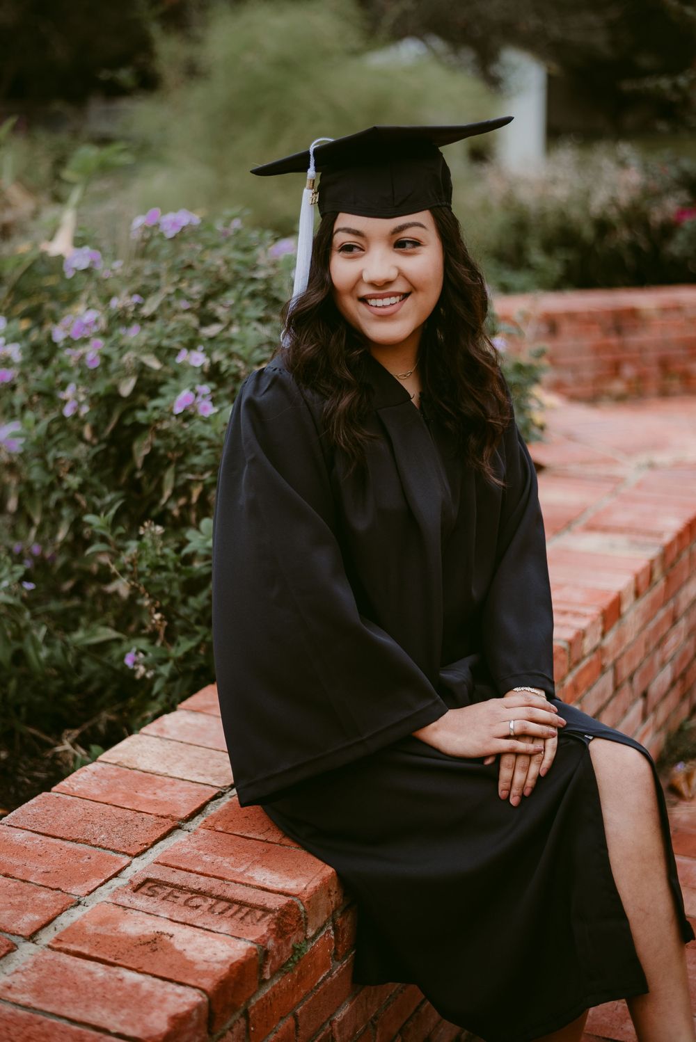 Graduate in black cap and gown poses on brick ledge with purple flowers in background.