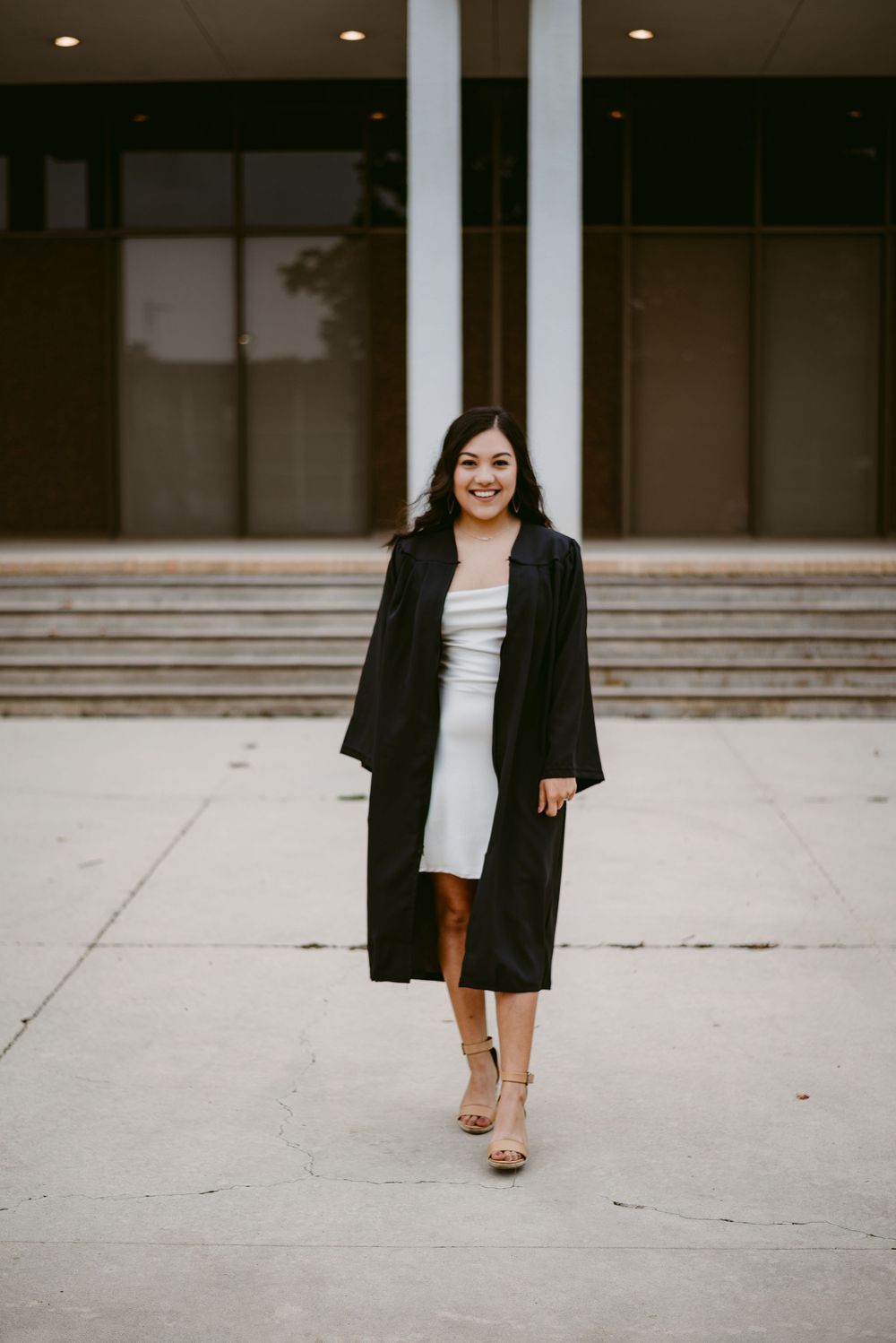 Graduate in academic dress walks confidently outside modern building with concrete steps.