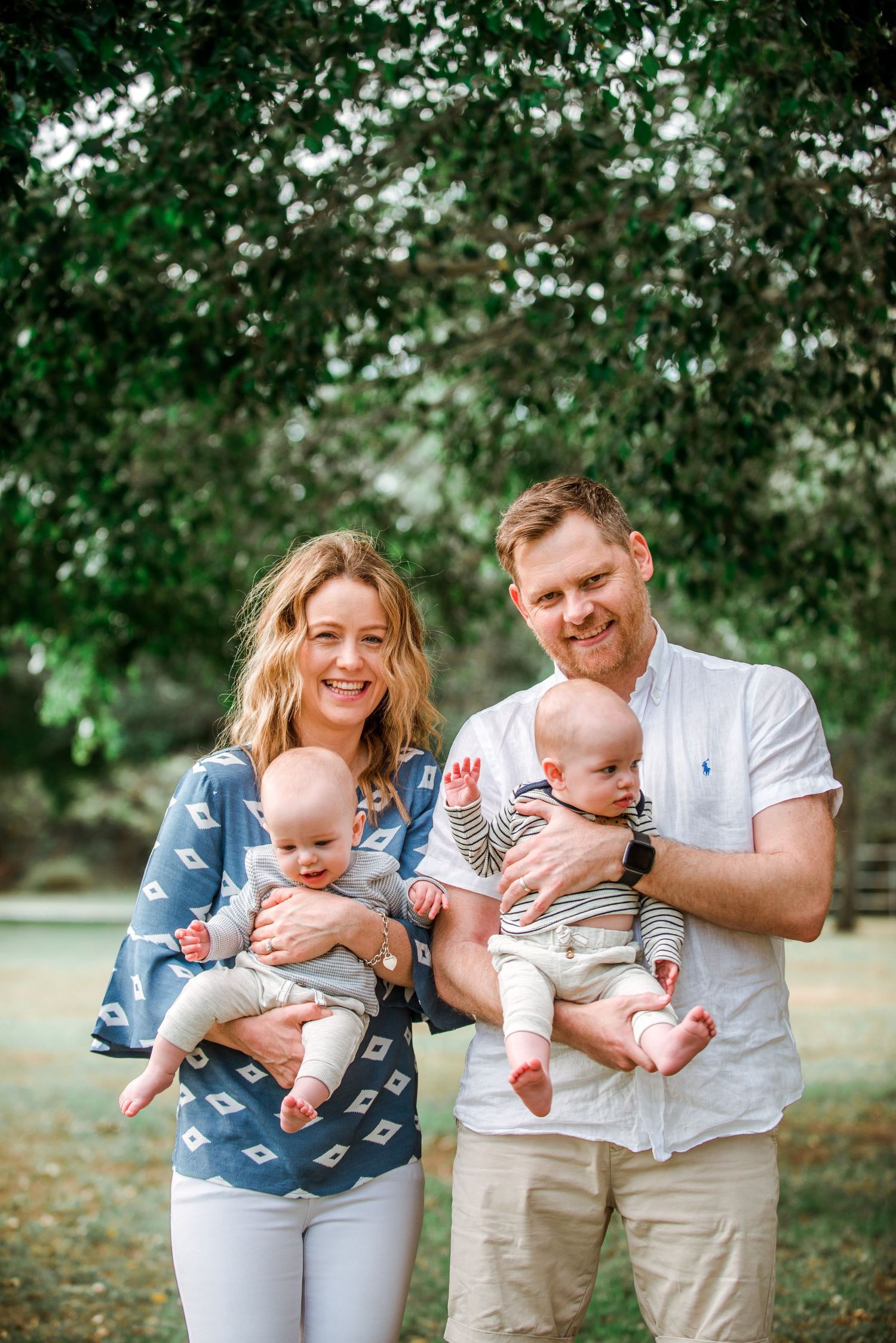 Joyful parents holding twin infants during outdoor family photo session with leafy background.