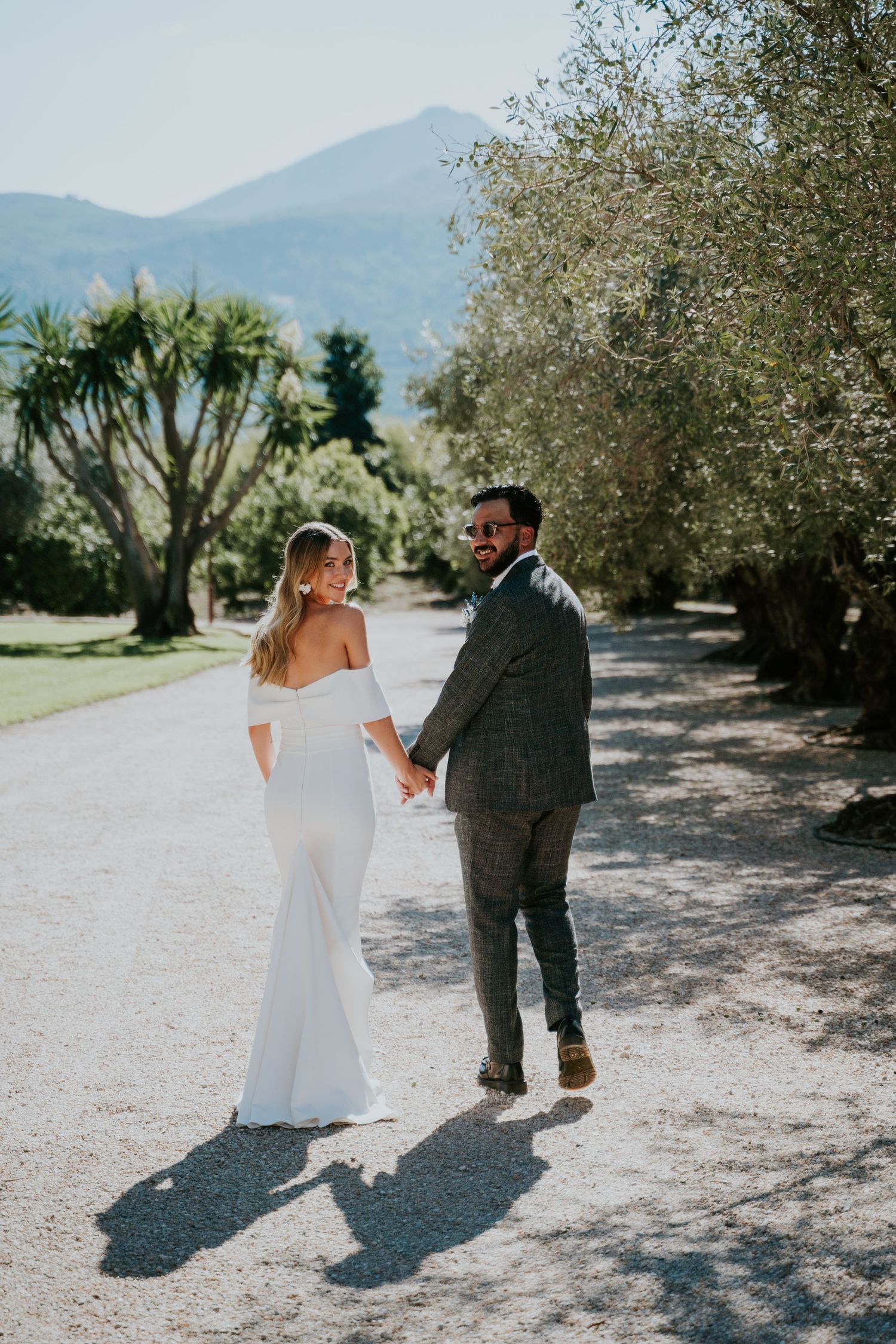 A couple in wedding attire walking hand in hand down a tree-lined path with mountains in the background.