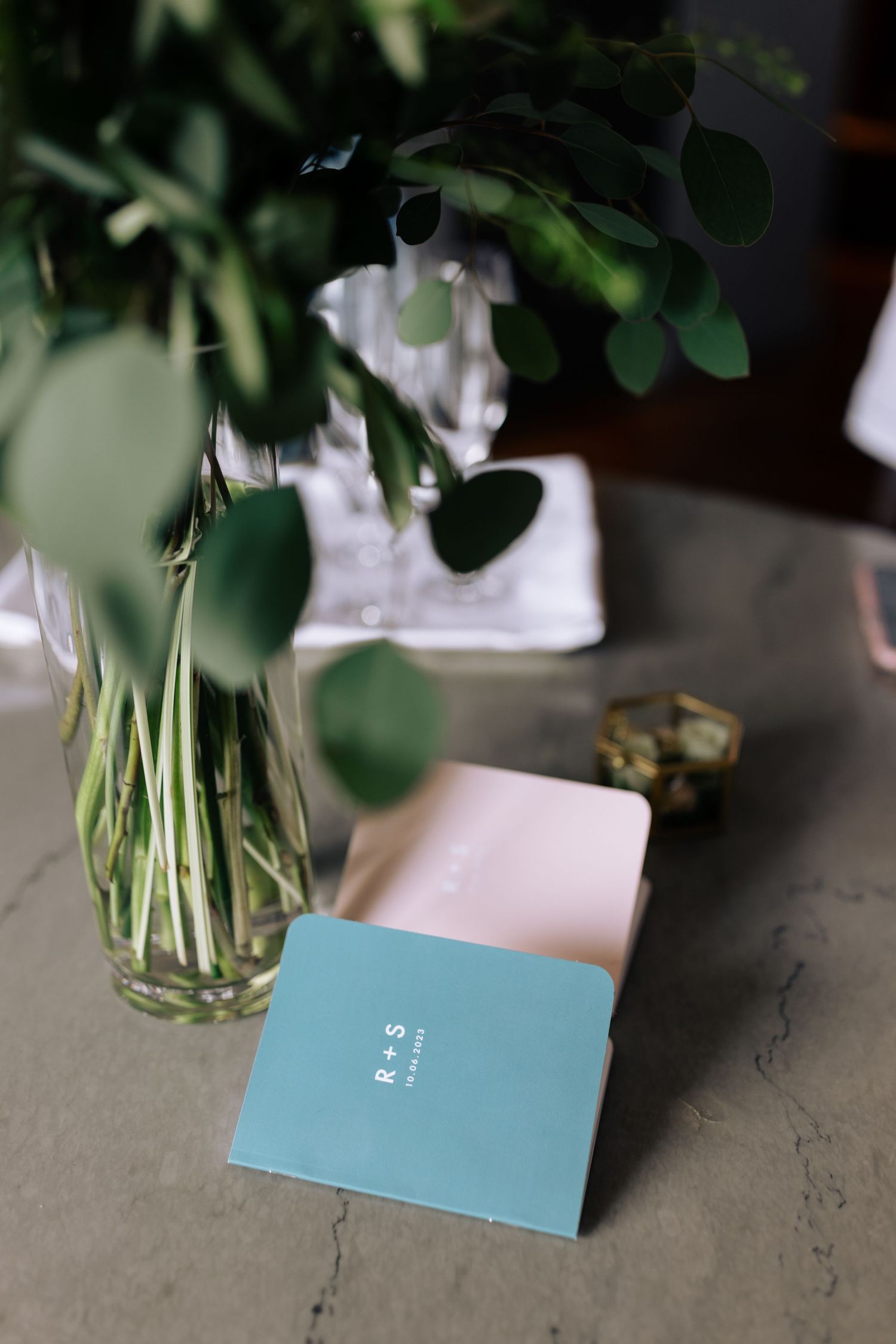 A turquoise book rests on a table next to a vase with greenery.