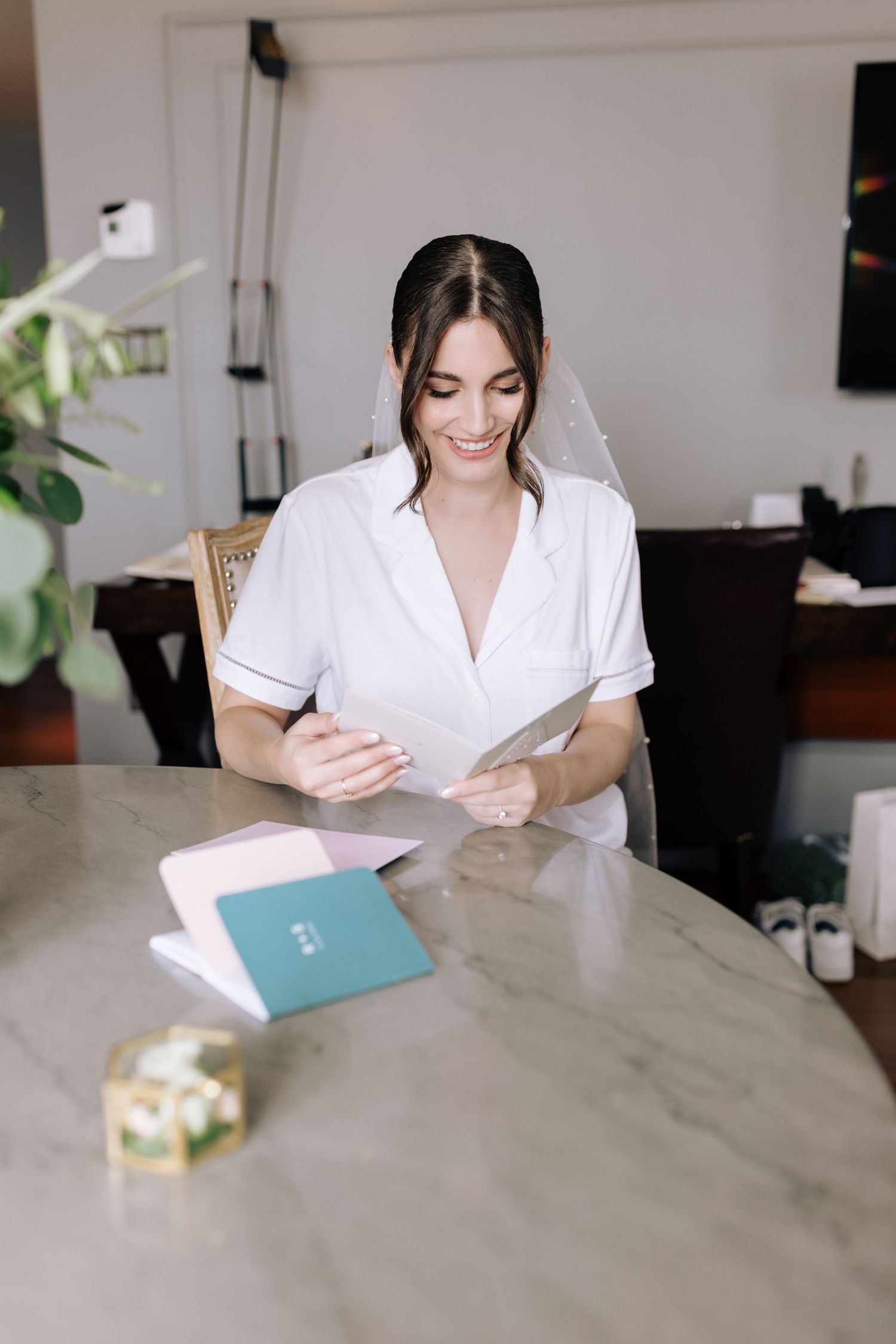 Bride in white robe reads letter at marble table during wedding morning preparations.