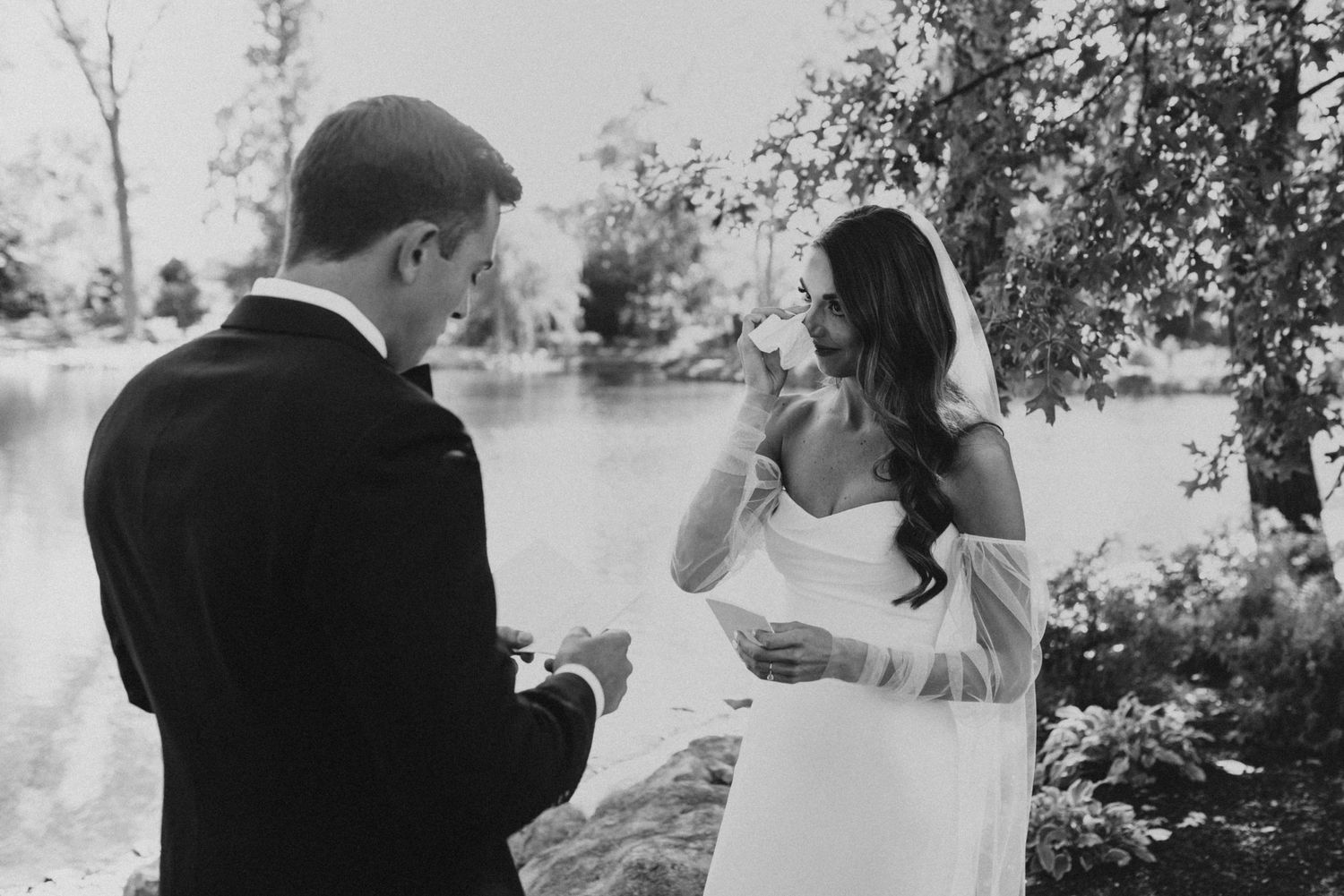 A black and white wedding photo capturing a couple's first look reaction by a lakeside.