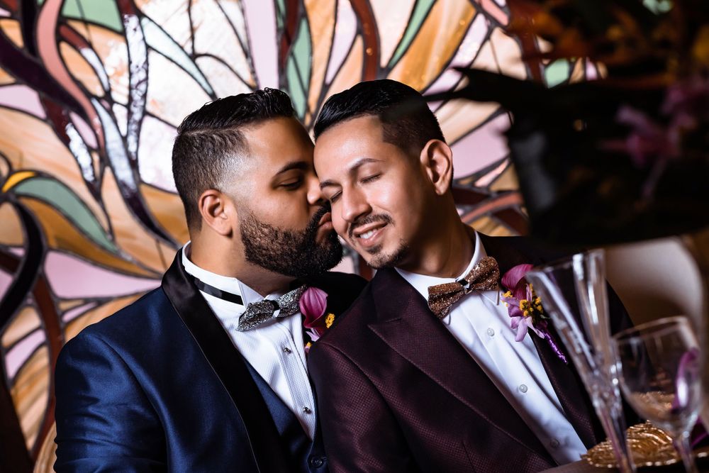 Two people in tuxedos share an intimate moment at a wedding with decorative plants in the background.