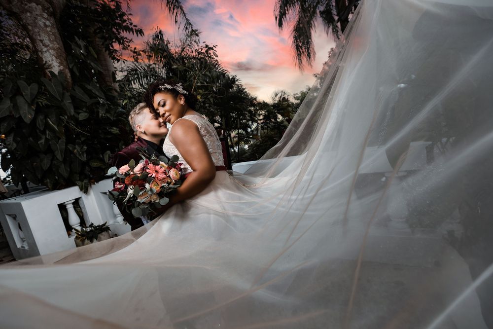 Artistic wedding photo at sunset with flowing veil and palm trees in background.