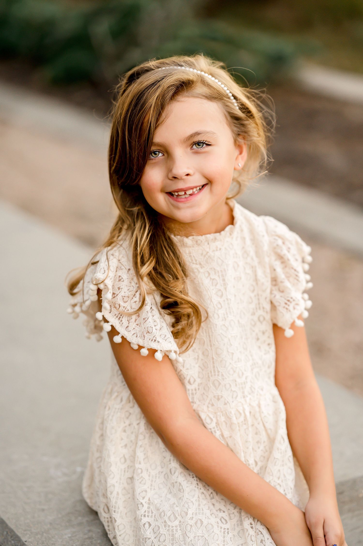 A child in a white lace dress smiles while posing outdoors.