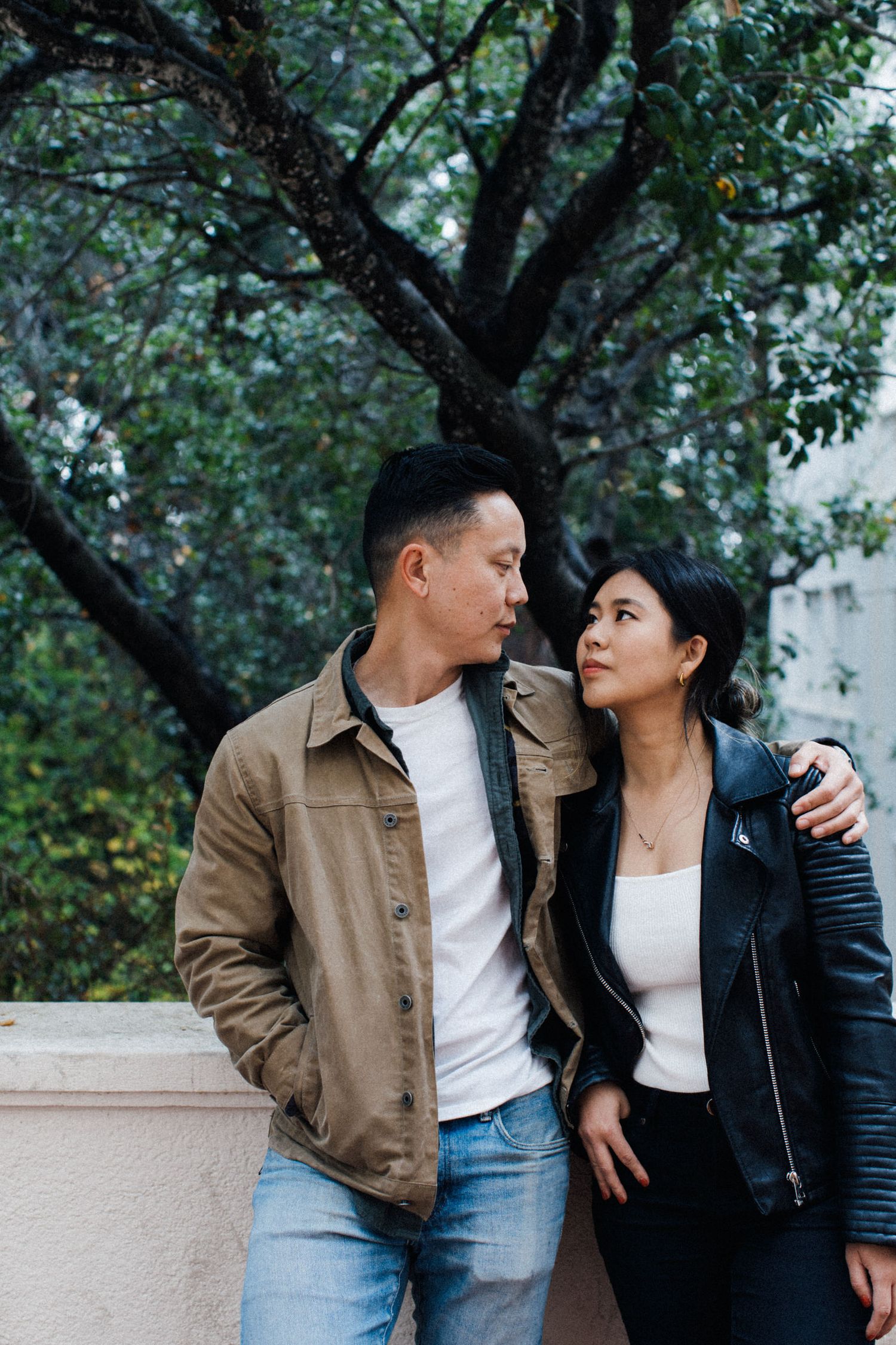 Stylish couple in leather and denim jackets sharing a romantic moment under a leafy tree.
