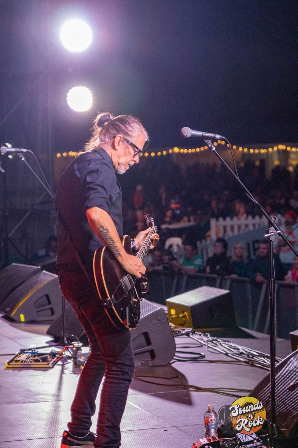 Guitarist playing on illuminated stage with string lights visible in background.