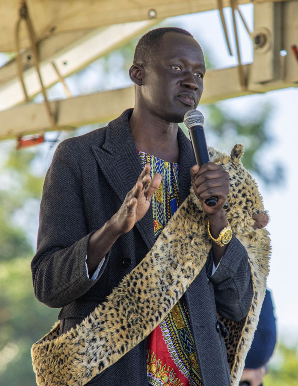 Speaker in dark jacket addresses audience while holding decorative leopard print sash on outdoor stage.