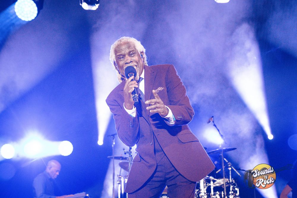 Concert performer in a burgundy suit sings passionately on stage with dramatic blue lighting effects.