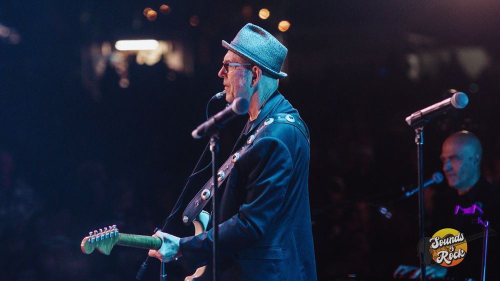 Performer in blue hat playing guitar on stage with concert lighting.