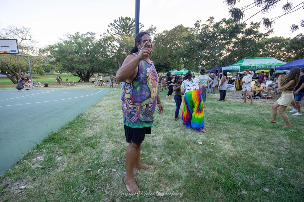 Person wearing colorful tie-dye shirt at outdoor festival with rainbow flag visible.