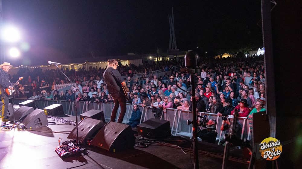 Large outdoor concert audience viewing performance from ground level with stage lights illuminating scene.