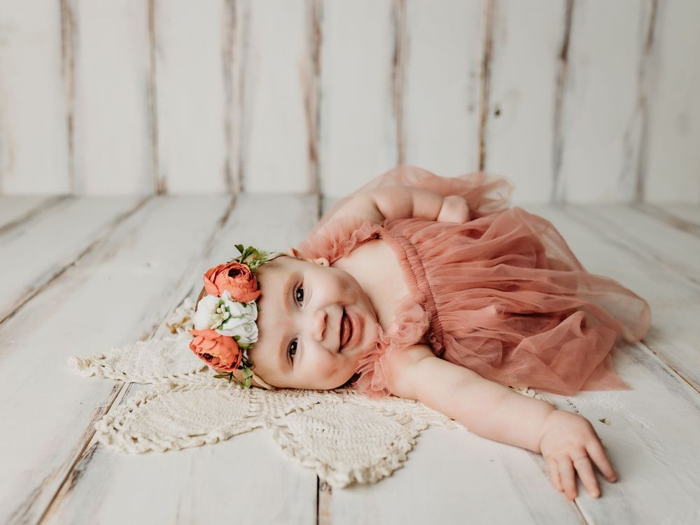 Happy baby wearing coral dress and floral crown laying on lace doily.
