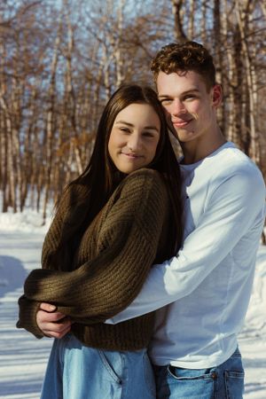 In a winter wooded area, a man hugs a woman from behind, her hands on top of his, and they both smile at the camera.