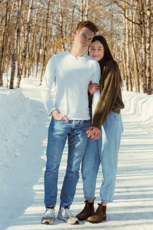 A couple in a winter wooded area stand facing the camera. The woman stands behind the man, wraps one arm around his, and holds hands.