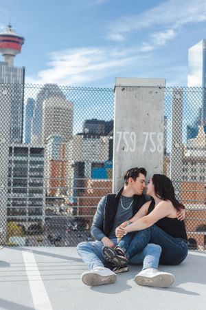 A couple sit on the ground of a parkade with the women's legs in the man's lap and they stare at each other. Calgary tower is in the back.
