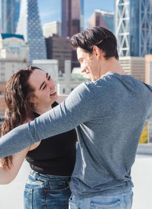A couple hold hands outstretched and stare at each other. City buildings in the background.