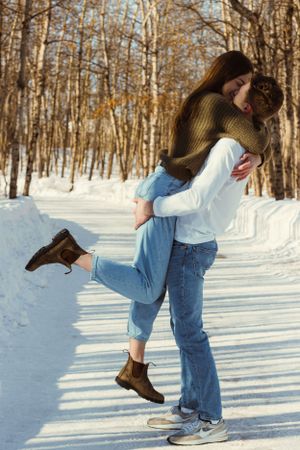 A couple kiss in a winter wooded area. The man is picking her up, the woman's arms are around his neck, and her leg is kicked up.