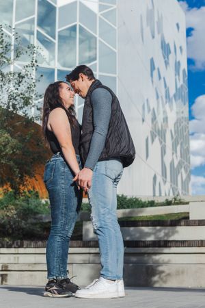 A couple stand facing each other, touching heads, and holding hands in front of a building.
