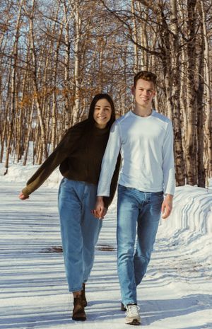 A couple in a winter wooded area walk towards the camera holding hands. The woman is leaning into the man.