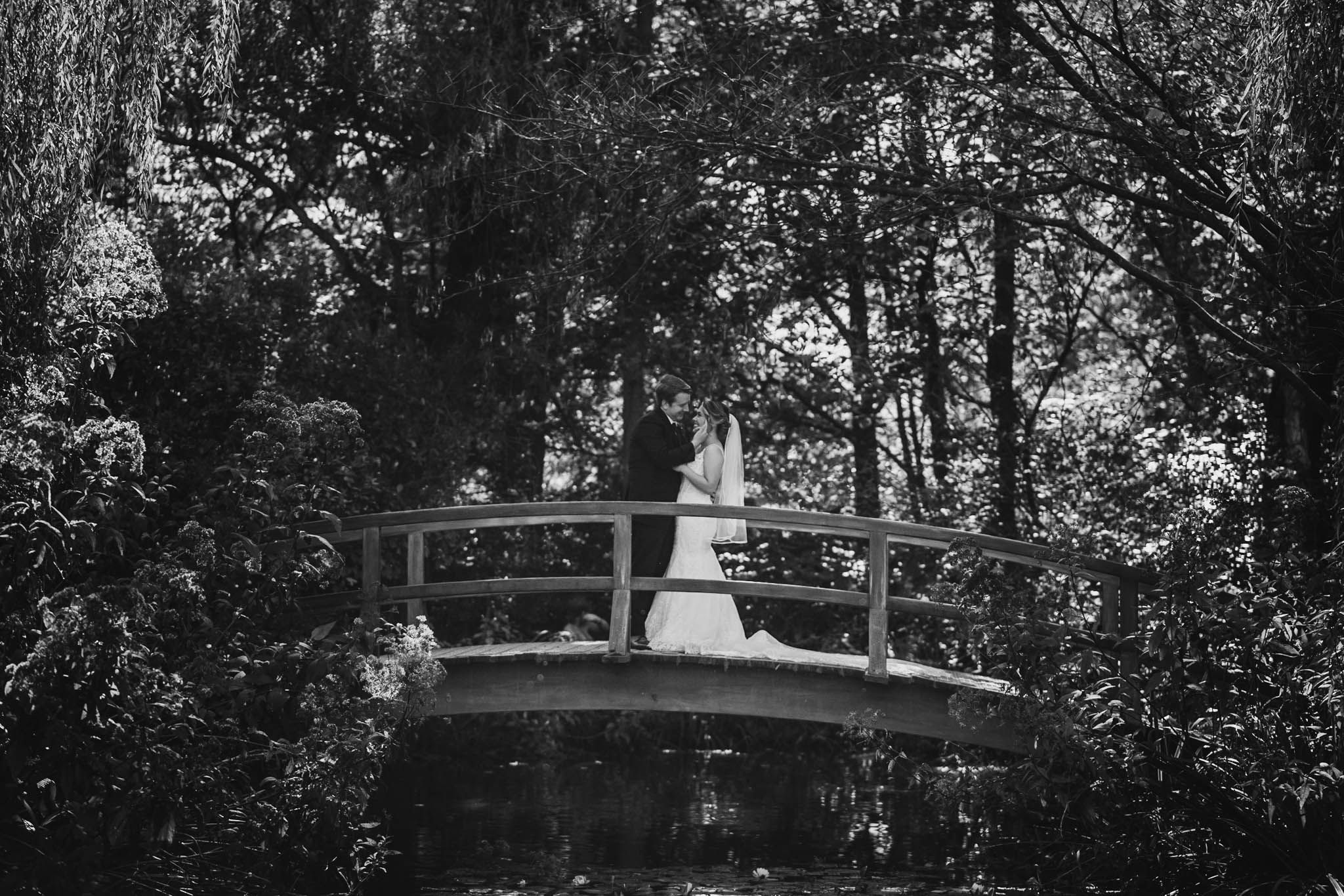 Black and white photo of newlyweds on a garden bridge with trees in background.