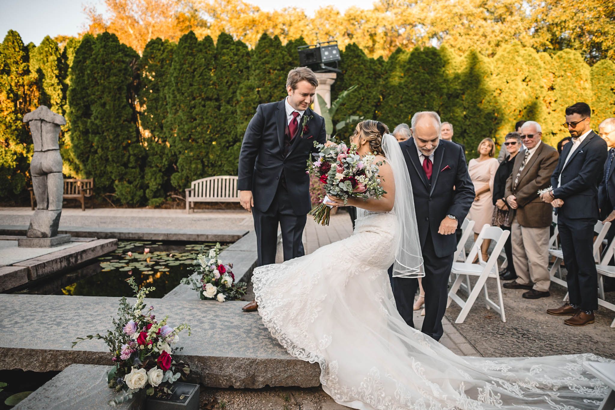Golden sunset light illuminates an outdoor garden wedding ceremony with guests seated.